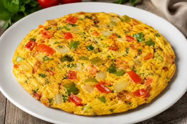 Fotografía de una tortilla de huevo con vegetales servida en un plato circular color blanco sobre una mesa de madera.